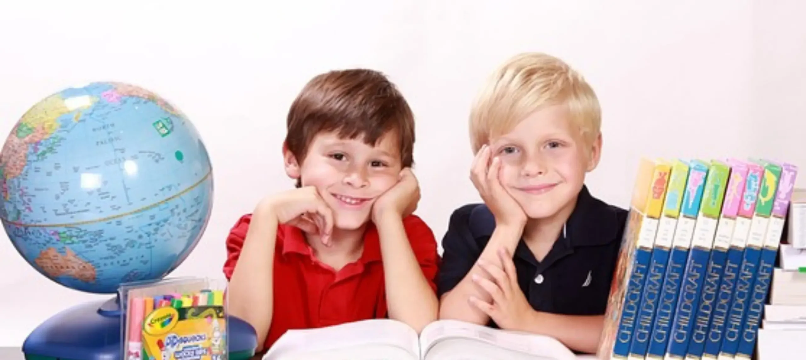 Intelligenztest bei Kindern Zwei Jungs sitzen zwischen Büchern und einem Globus und lächeln in die Kamera