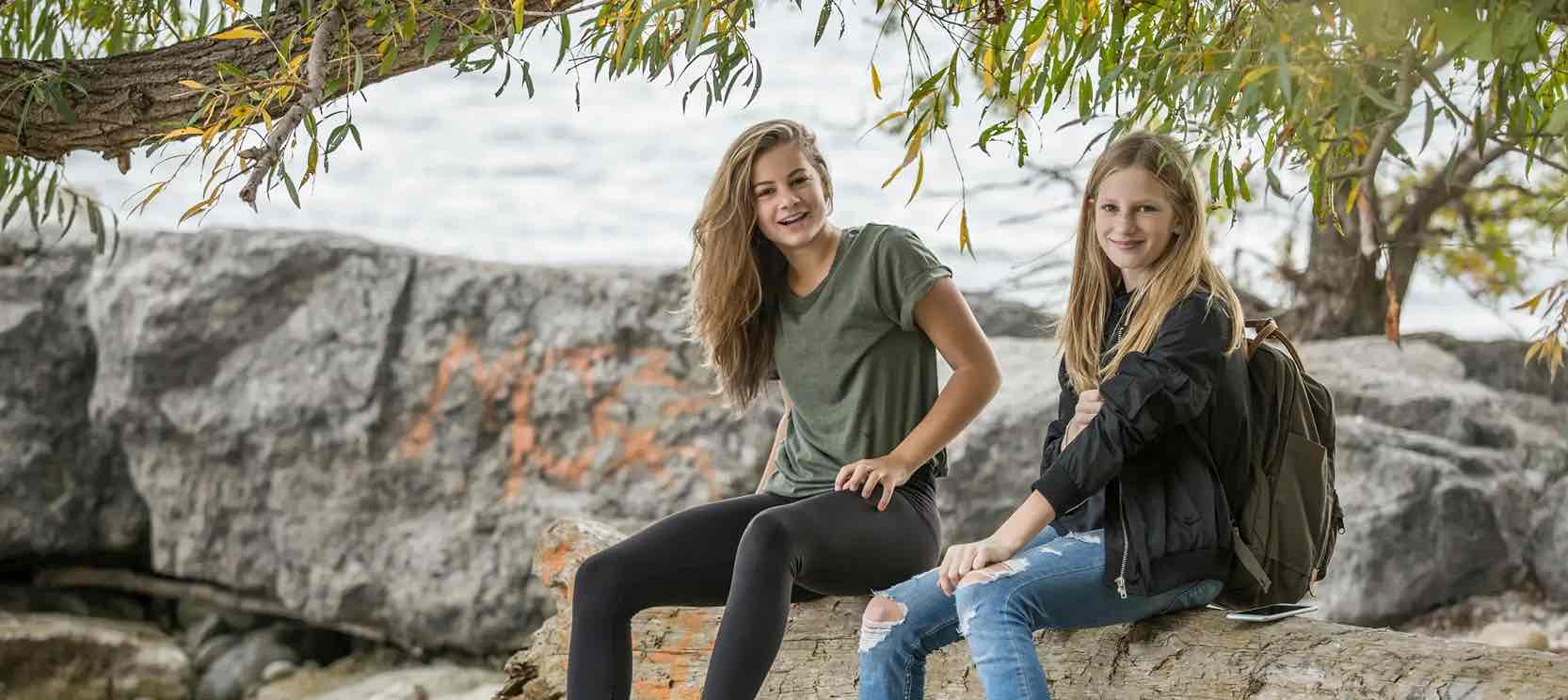 Pubertät bei Mädchen Zwei Mädchen sitzen auf einem Baumstamm in der Nähe eines Gewässers