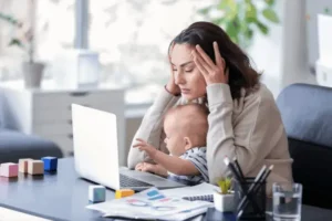 Eine Mutter sitzt vor eine Laptop und hat ein Baby auf ihrem Schoß. Ihre Hände hält sie and ihre Schläfe und sie sieht gestresst und überfordert aus.